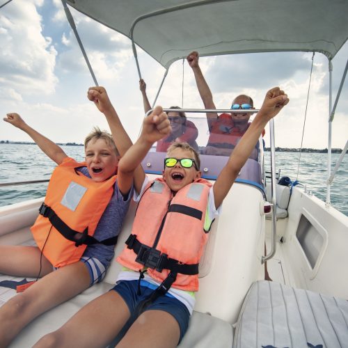 Family enjoying riding a boat on Lake Garda Two excited children wearing life jackets raise their arms and cheer while sitting on a speedboat, with two adults smiling behind them; the boat is on a lake under a partly cloudy sky.