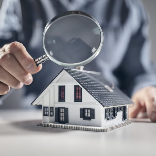 House model with man holding magnifying glass home inspection or searching for a house A person holds a magnifying glass over a small model house, symbolizing close inspection or evaluation of real estate property.