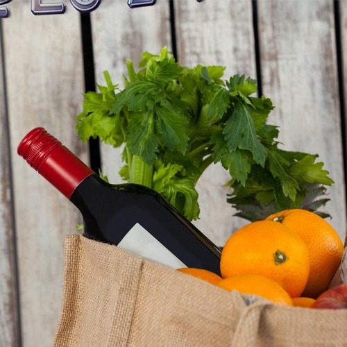 Groceries A burlap bag filled with oranges, celery, and a bottle of red wine with a blank label, set against a rustic wooden background.